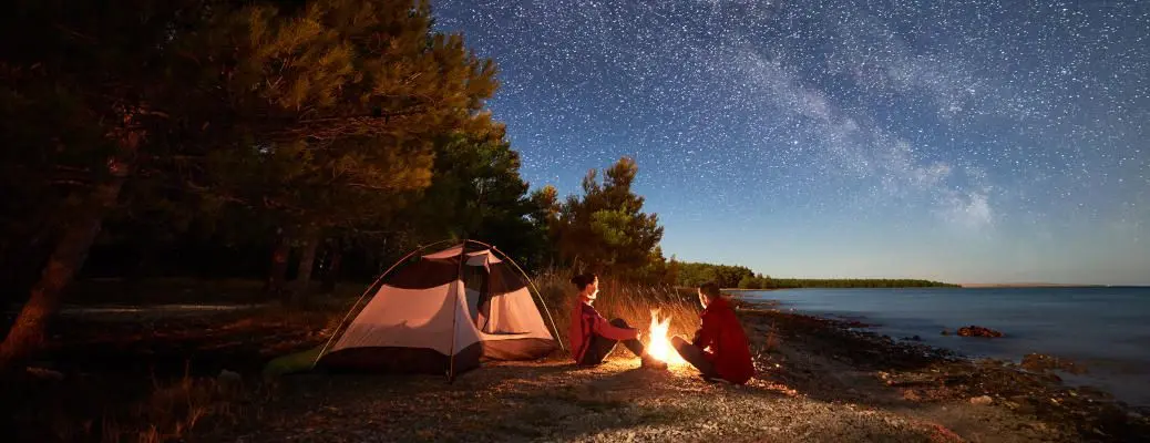 Young couple man and woman having rest at tourist tent and burning campfire on sea shore near forest