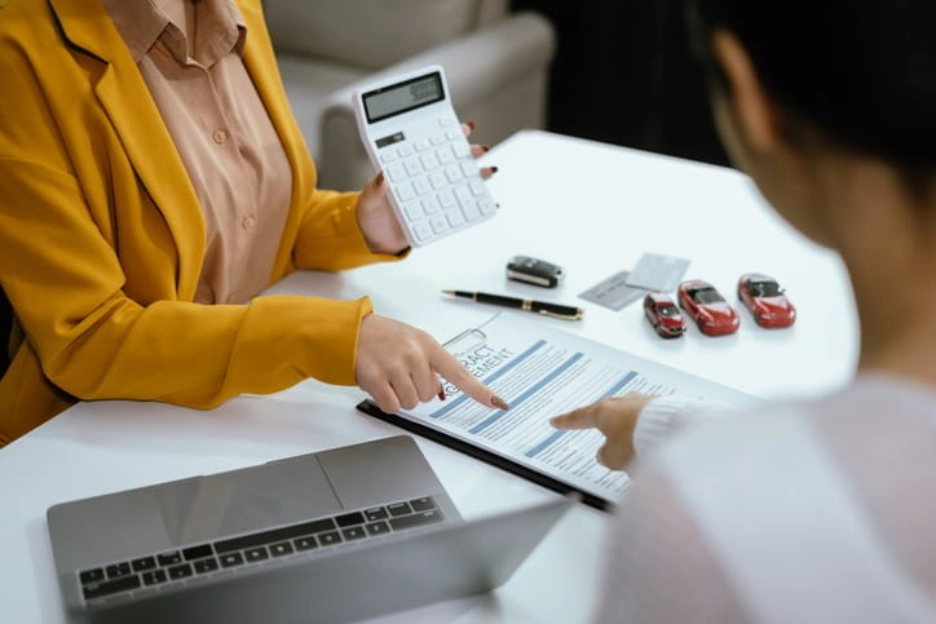 Two people reviewing a financial document at a desk, one holding a calculator while pointing to a contract, with a laptop and small model cars nearby.