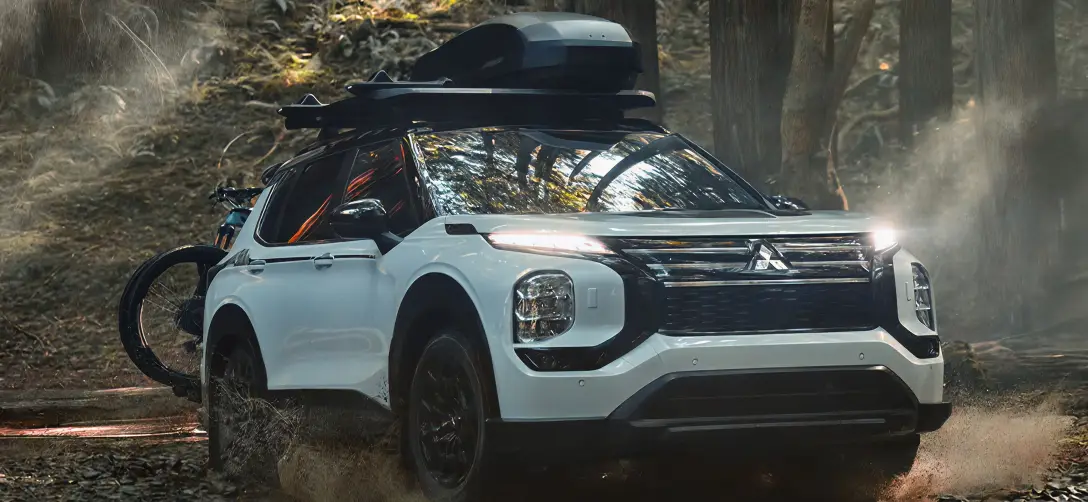 White Mitsubishi Outlander with roof cargo and bikes driving through a wooded trail.