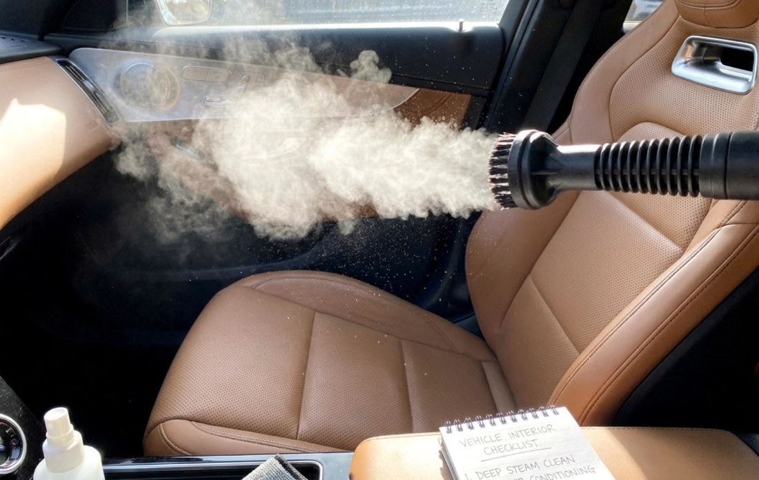 Steam cleaner nozzle releasing steam inside a vehicle interior with tan leather seats, highlighting deep interior detailing.