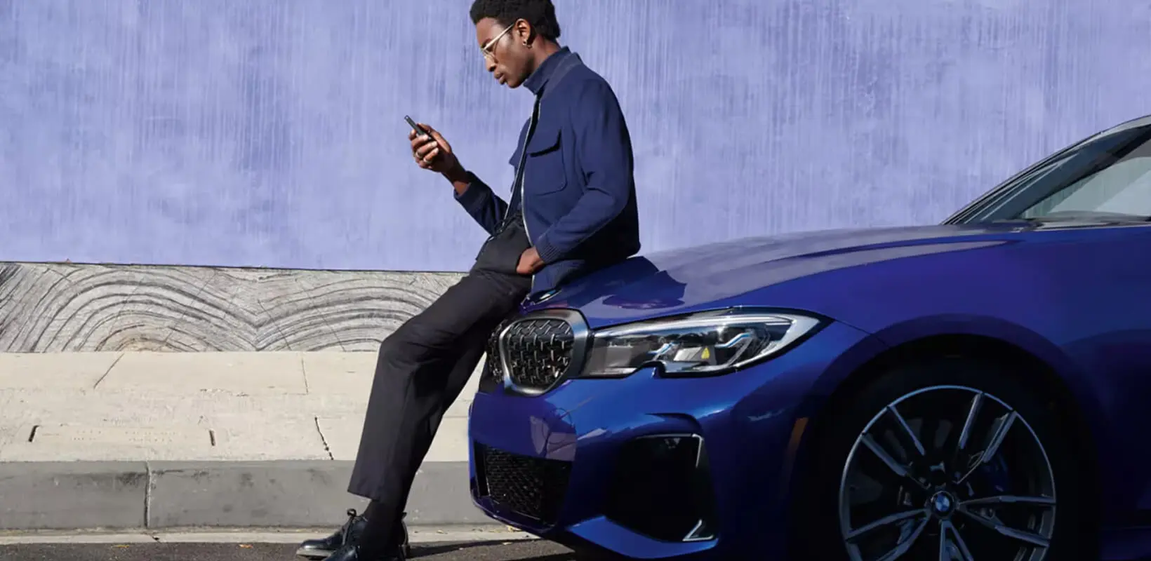 A man leans on the hood of his BMW looking at his phone.
