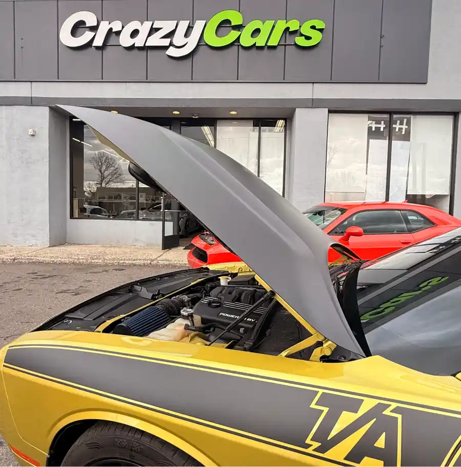Yellow car with the hood open parked in front of the Crazy Cars dealership building.