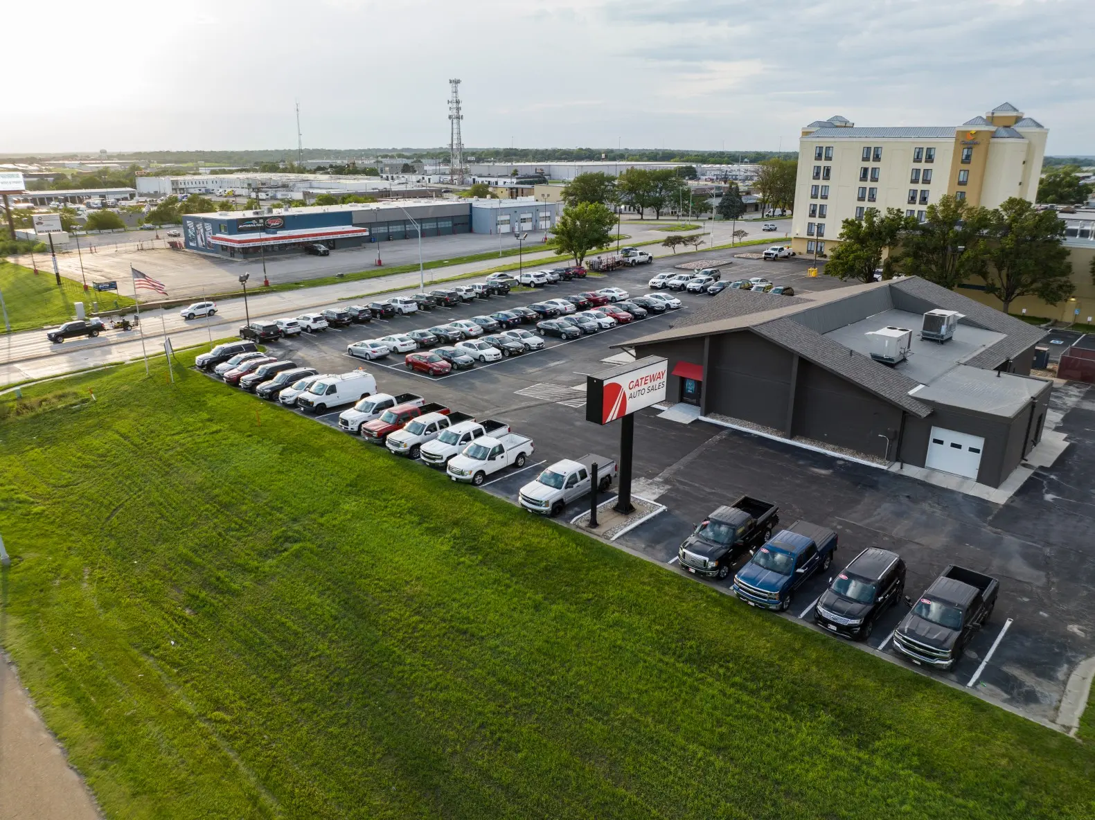 Premier used car dealership lot in Papillion, NE at golden hour with polished sedans, SUVs, pickups, and crossovers. Sunlight highlights authentic vehicle details, blurred price stickers, waving American flags, modern building, and leafy Nebraska trees under a partly cloudy sky.