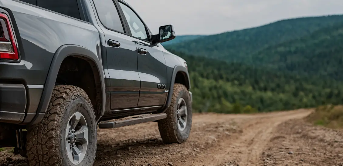 Close-up of a pickup truck driving on a dirt road with mountain landscape.