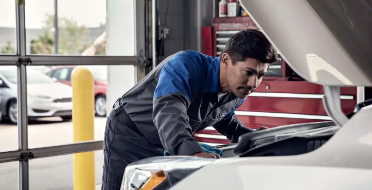 Ford technician inspecting under the hood of a white vehicle in a service bay