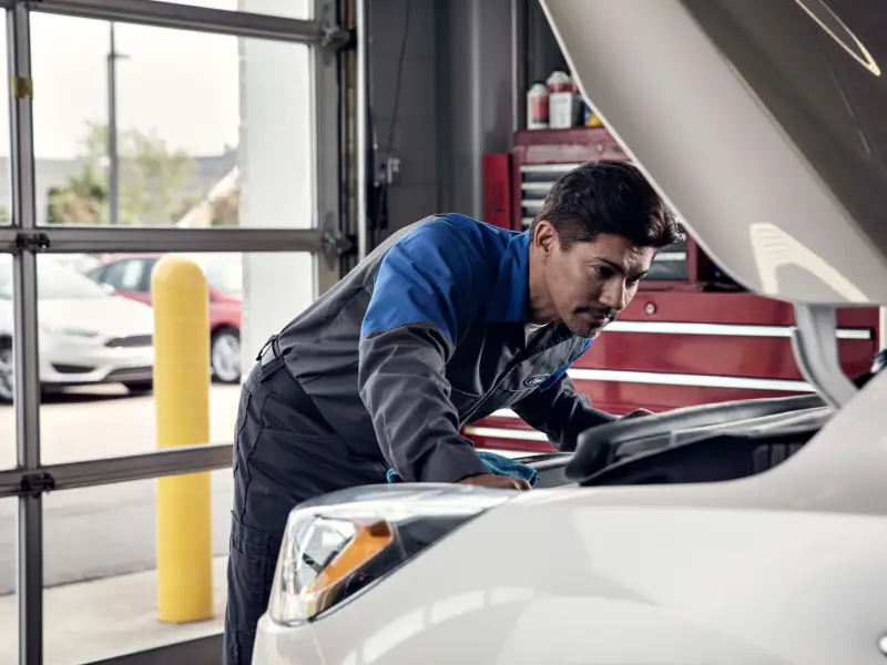 Ford technician inspecting under the hood of a white vehicle in a service bay