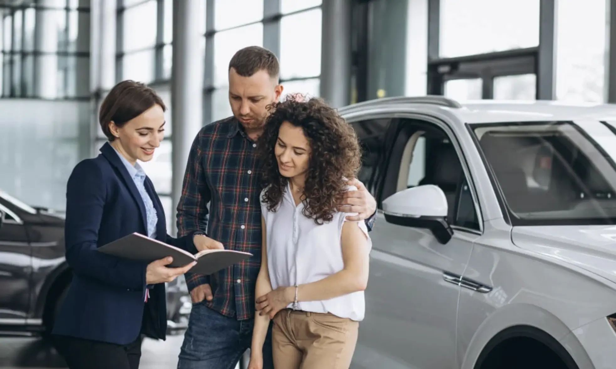 Couple reviewing paperwork with a saleswoman next to a silver SUV inside a dealership showroom