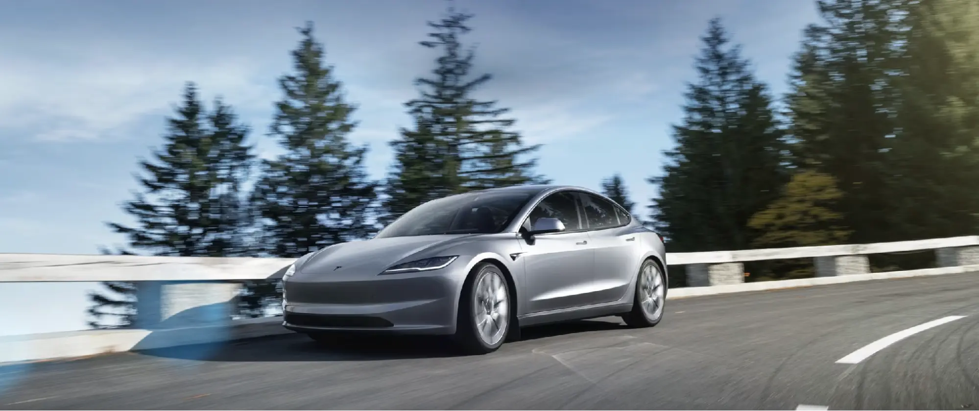 Silver pre-owned Tesla Model 3 driving on a winding road lined with pine trees