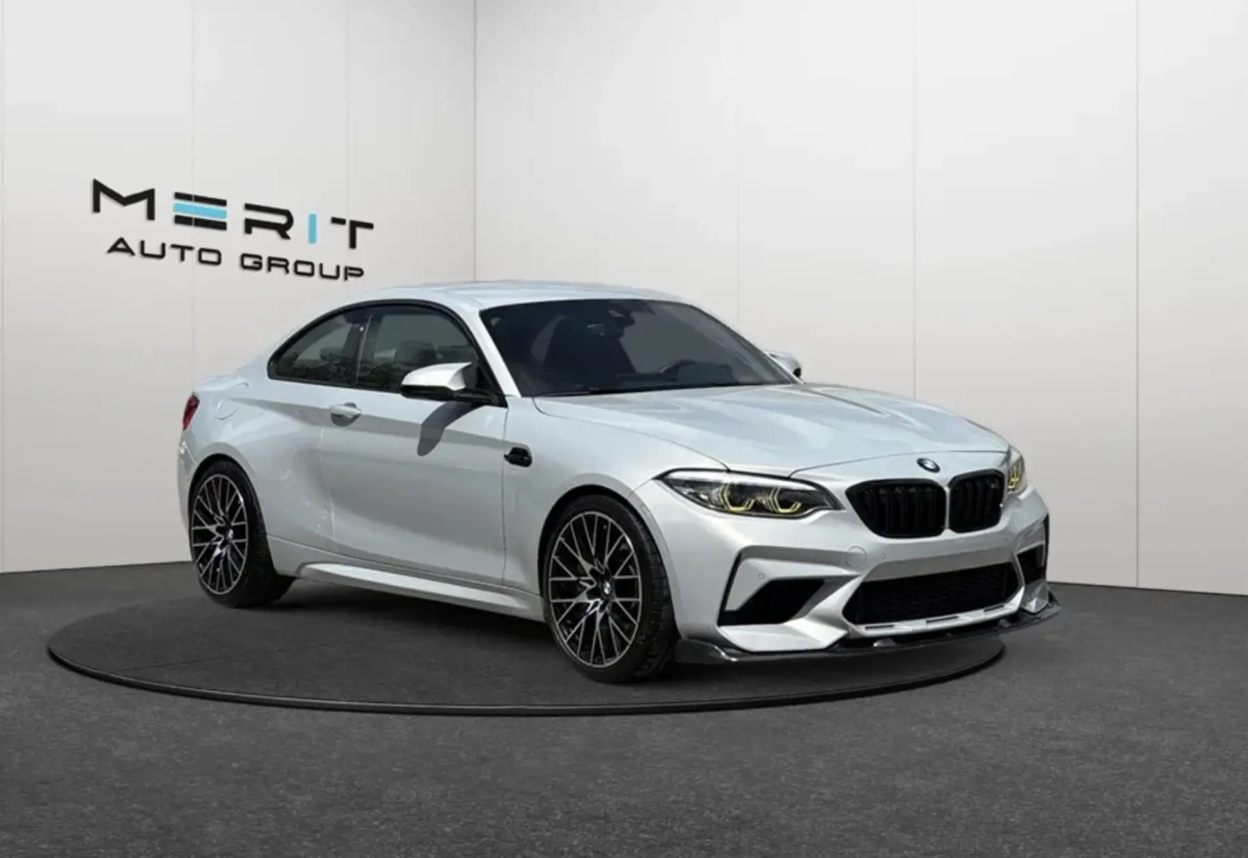 Silver BMW M2 Competition coupe displayed on a turntable in front of the Merit Auto Group showroom logo