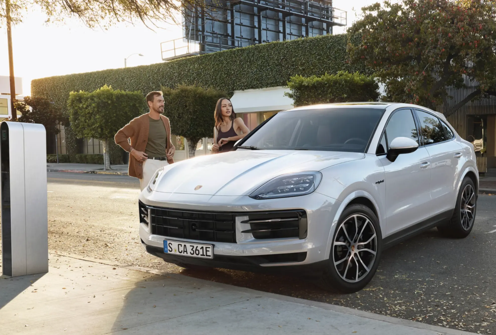 Silver Porsche Cayenne Coupe parked on a city street at sunset with a couple standing beside it.