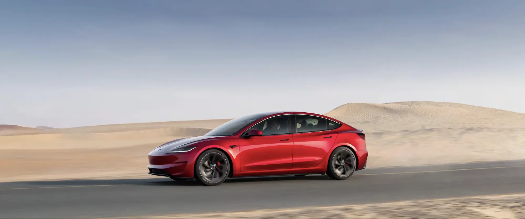 Red used Tesla Model 3 driving on a desert road past sand dunes