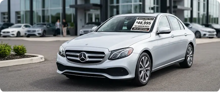 Silver Mercedes-Benz E-Class with a wholesale price window sticker in the Merit Auto Group lot, front three-quarter view