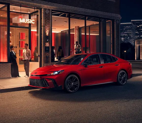 Red Toyota Camry parked outside a storefront at dusk, front three-quarter view