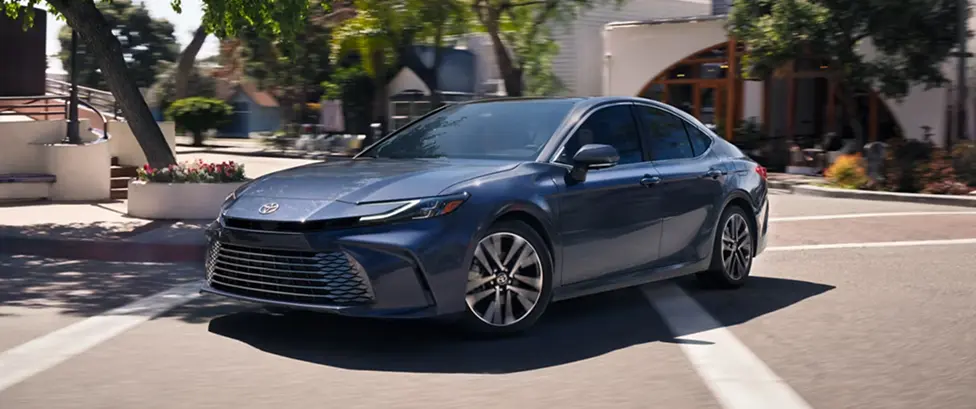 Blue-gray Toyota Camry driving down a tree-lined street, front three-quarter view