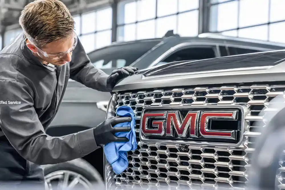 Technician wearing safety glasses and gloves polishing the front grille of a GMC vehicle in a service shop.