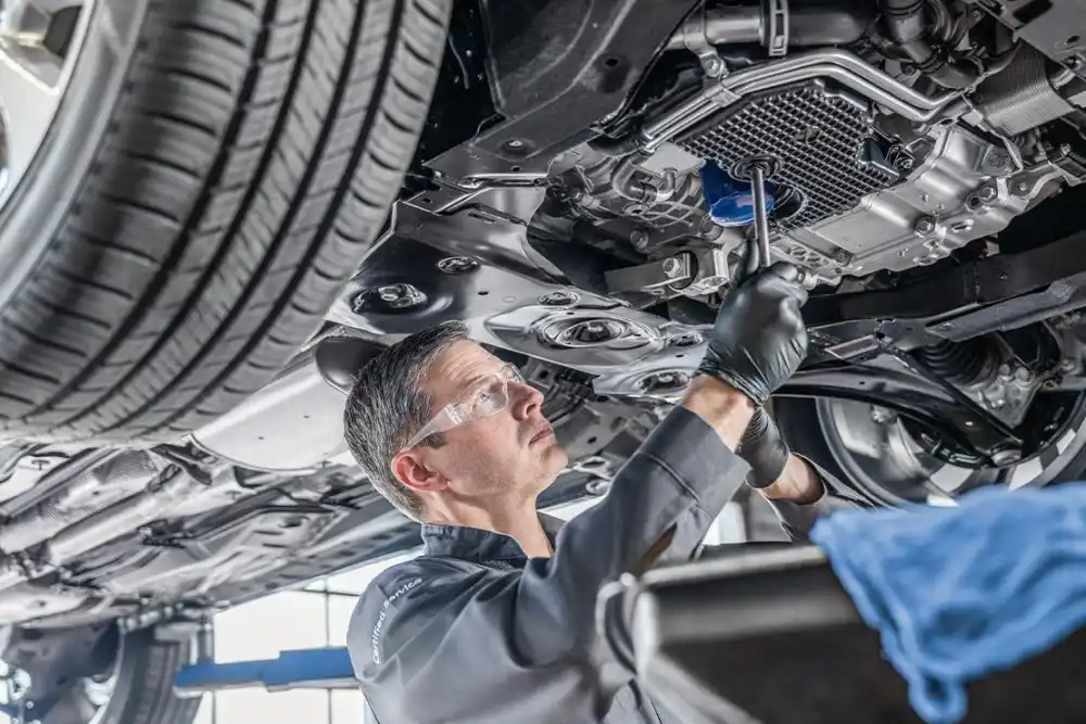 Technician wearing safety glasses and gloves inspecting the underside of a vehicle raised on a lift.