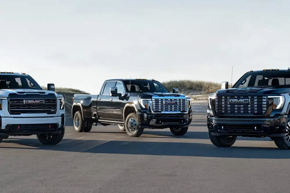 Three GMC pickup trucks in white and black lined up on a paved lot near a coastal dune landscape.