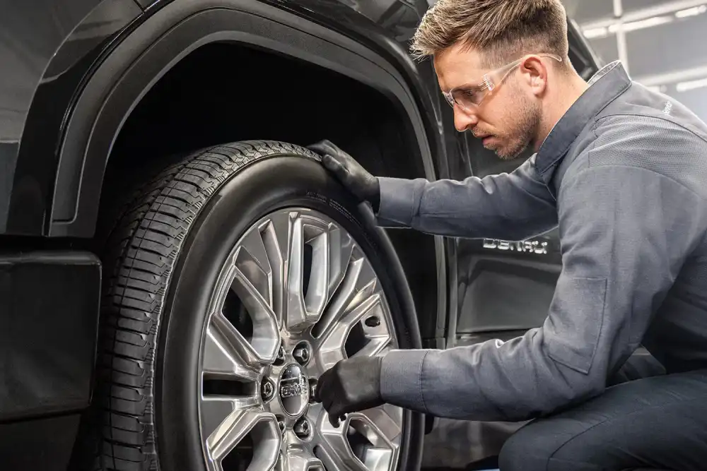 Technician wearing safety glasses inspecting a wheel and tire on a GMC vehicle in a service bay.