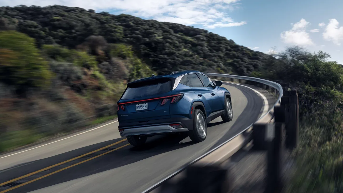 Blue 2026 Hyundai Tucson driving on a winding hillside road, rear three-quarter view