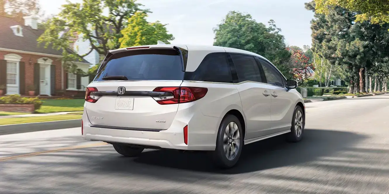 White 2027 Honda Odyssey Elite driving on a suburban street, rear three-quarter view