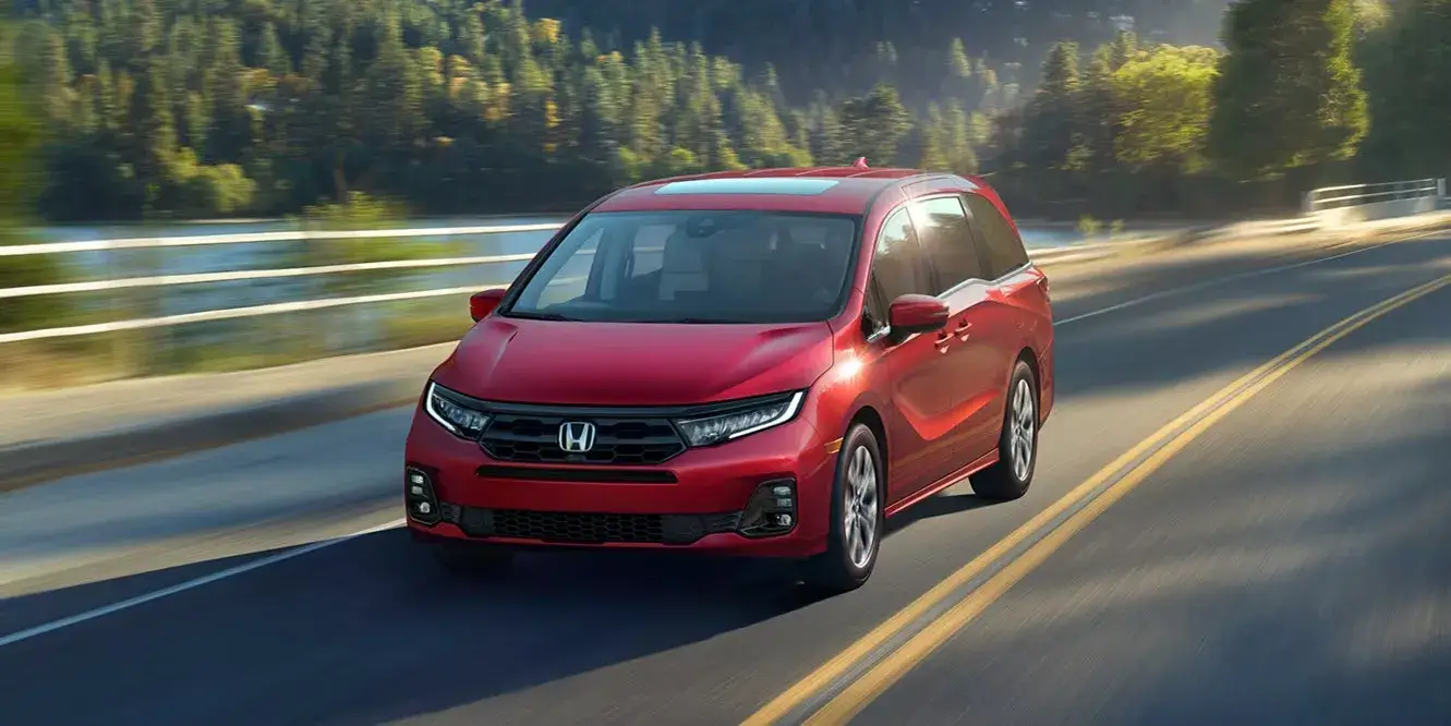 2027 Honda Odyssey Elite in Radiant Red Metallic II driving on a lakeside road, front view