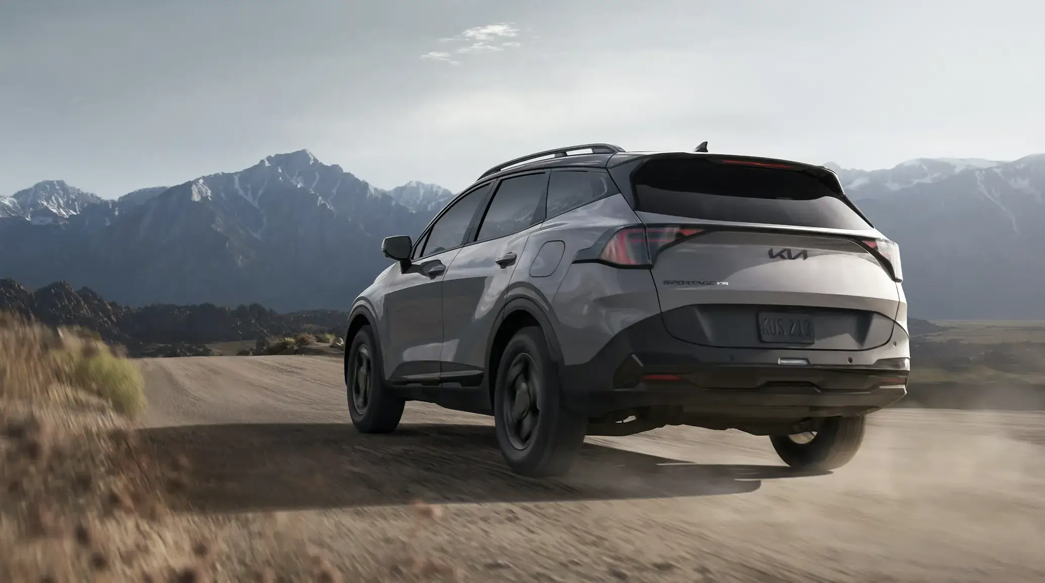 Gray 2027 Kia Sportage driving on a desert dirt road, rear three-quarter view