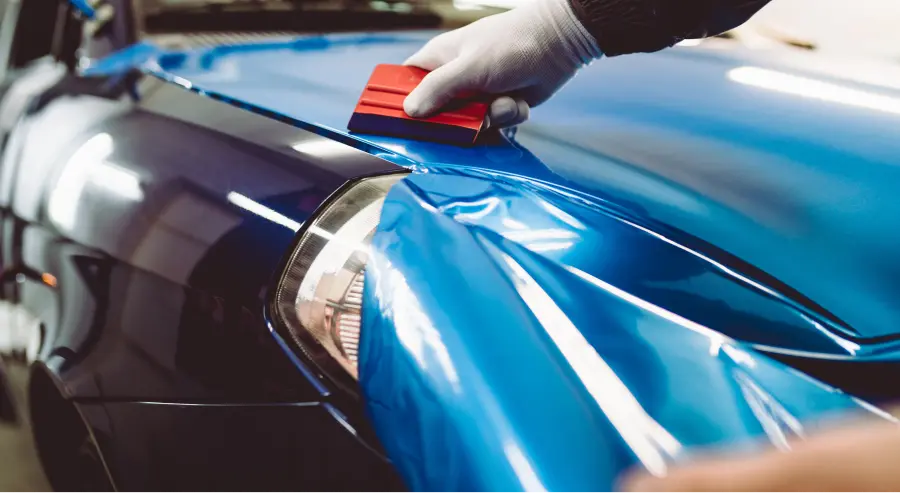 Technician smoothing blue vinyl wrap over a car's hood and front fender